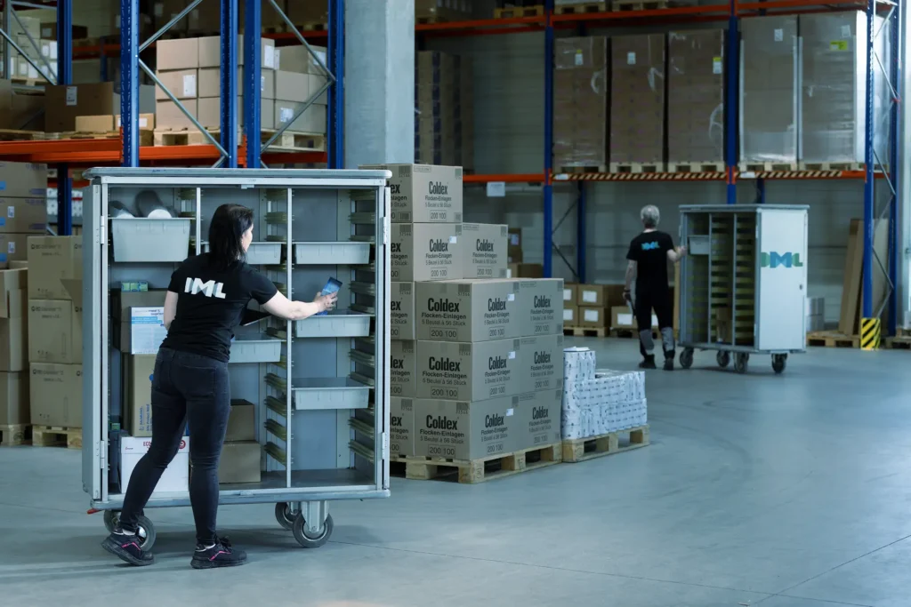 Zwei IML Mitarbeiter:innen sind in der Lagerhalle unterwegs, um Bestellungen von medizinischem Material in ihren Trolleys zusammenzustellen.